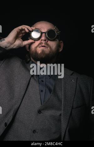 Charismatischer Mann in klassischem Anzug und Steampunk-Brille auf schwarzem Hintergrund stilvolles Porträt Stockfoto