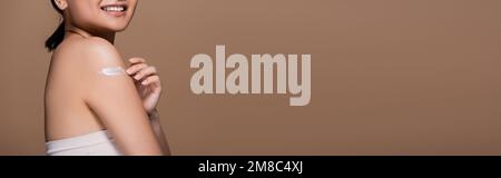 Zugeschnittene Ansicht einer positiven Frau oben, die Kosmetikcreme auf der Schulter aufträgt, isoliert auf einem braunen Banner, Stockbild Stockfoto