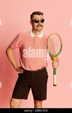 Fröhlich. Porträts eines gutaussehenden charismatischen Mannes in stilvoller Kleidung mit Tennisschläger auf pinkfarbenem Studiohintergrund. Das Konzept von Mode, Sport Stockfoto