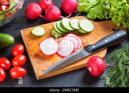 Geschnittene Zutaten für den Salat auf dem Schneidebrett mit Messer. Frisches Gemüse auf schwarzem Schieferhintergrund. Draufsicht. Stockfoto