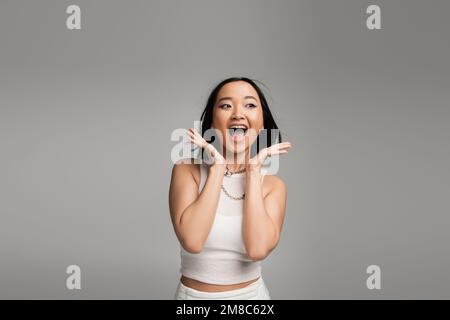 Aufgeregte asiatische Frau in weißem Tanktop, die Hände nahe dem Gesicht hält und schreit isoliert auf grauem, Stock Image Stockfoto