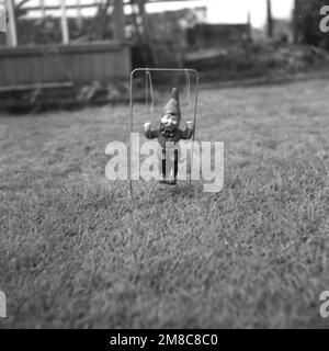 1950er, historisch, ein kleiner Gartenzwerg auf Schaukel saß auf einem Rasen, England, Großbritannien. Stockfoto