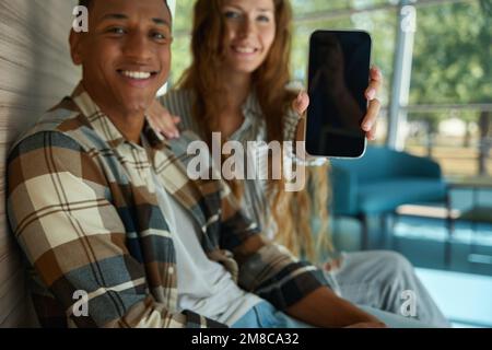 Glückliche Frau, die das Telefon zeigt und neben ihrem männlichen Kollegen sitzt Stockfoto