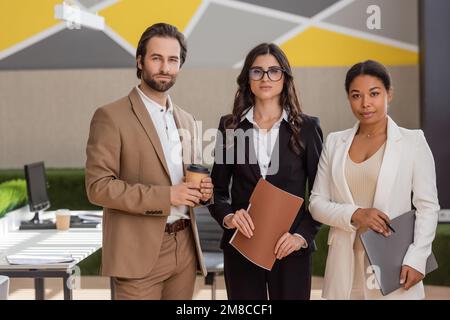 Multiethnische Geschäftsleute mit Laptop und Kaffee, die im Büro vor die Kamera schauen, Bestandsbild Stockfoto