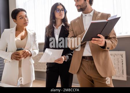 Der junge Manager zeigte der multikulturellen Geschäftsfrau, die mit Papier und einem Getränk zum Mitnehmen stand, ein Bild von der Börse Stockfoto