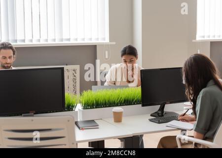 Konzentrierte, multiethnische Geschäftsleute, die in der Nähe von Monitoren mit leerem Bildschirm in modernen Büros arbeiten, Aktienbild Stockfoto
