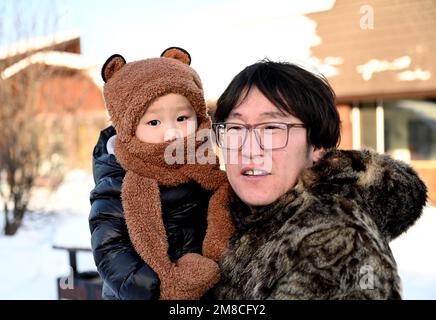 (230113) -- GENHE, 13. Januar 2023 (Xinhua) -- Dieses Foto wurde am 13. Januar 2023 aufgenommen und zeigt Juele mit seinem Kind in einer neuen Umzugsgemeinde in Aoluguya Ewenki Township in Genhe City, Nordchinas innere Mongolei Autonome Region. Alle zwei oder drei Tage fährt Juele Bulituotian zum Jinhe-Waldgebiet etwa 80 Kilometer von Genhe City entfernt. Dort wird der 39-jährige Aoluguya Ewenki-Hirte, der einen traditionellen Pelzmantel trägt, sein forschendes Rentier finden. Anstatt Rentiere in einer Scheune zu halten, lassen die Ewenki-Hirten die Tiere im Wald leben und überprüfen sie alle paar Tage. Juele Stockfoto