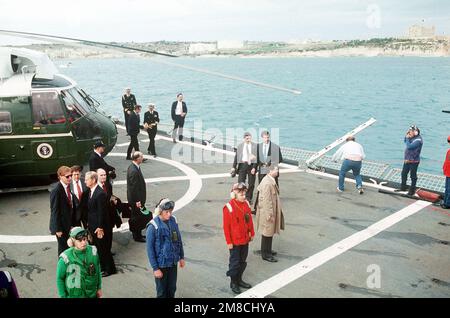 Hinter einer Reihe von Besatzungsmitgliedern des Marine Helicopter Squadron One (HMX-1) berät sich Außenminister James Baker mit Mitgliedern der Präsidentschaftspartei, während Präsident George H.W. Bush wird von einem Marineoffizier begrüßt. Bush besucht den geführten Raketenkreuzer USS BELKNAP (CG 26) vor seinem Gipfeltreffen im Dezember 2-3 mit dem sowjetischen Vorsitzenden Michail Gorbatschow. Basis: Marsaxlokk Bay Country: Malta (MLT) Stockfoto