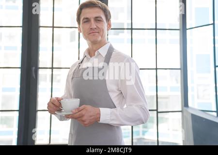 Ein Kellner in einer grauen Schürze hält eine Tasse Kaffee. Der Barista gibt eine Tasse heißen Kaffee in einem Café vor dem Hintergrund eines großen Fensters. Hochwertiges Foto Stockfoto