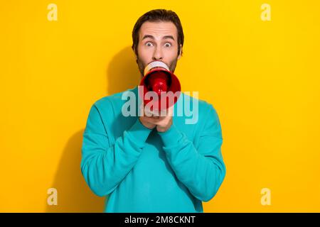 Foto eines beeindruckten, schockierten Mannes im blaugrünen Outfit, der ein Megafon mit isoliertem gelbem Hintergrund macht Stockfoto