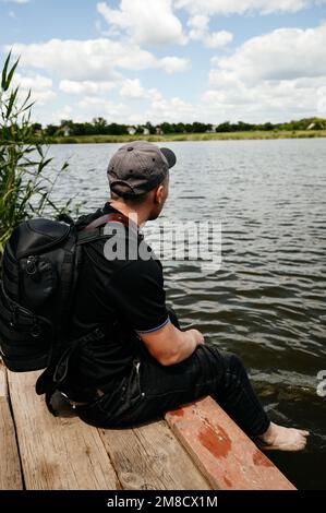 Ein Mann sitzt am Rand einer hölzernen Brücke in der Nähe eines Sees neben Schilf, einem tiefen und klaren Gewässer am Horizont. Stockfoto