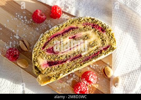 Grüne Pistazien-Baiser-Roulade-Scheibe mit Himbeermarmelade auf hellem Holzbretthintergrund in der Küche von oben. Stockfoto