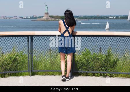 Eine Frau, die von der Gouverneursinsel auf die Freiheitsstatue auf ellis Island in New York City blickt. Stockfoto