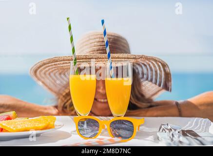 Nahaufnahme von Orangensaftgläsern und gelber Sonnenbrille, vor einer Frau mit Strohhut im Hintergrund unscharf. Blaues Meer im Hintergrund. Urlaub Stockfoto