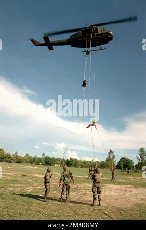 Angriffsleute stehen vor Ort als vier Mitglieder von 4. Milliarden, 22. Inf., 25. Inf. Bereit. Div (Licht), Abseilen von einem 53. Luftbataillon UH-1H Iroquois Helikopter während des Trainings als Teil der Kombination Thai/USA Übung Cobra Gold '90. Betreff Operation/Serie: COBRA GOLD '90 Basis: Chon Buri Land: Thailand (THA) Stockfoto