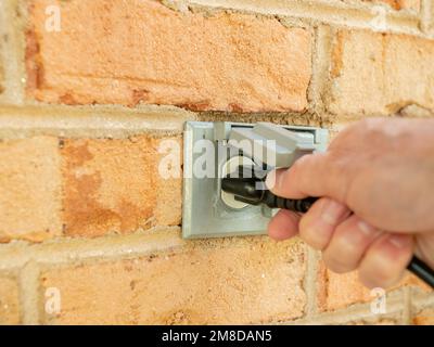 Mann, der ein Netzkabel einsteckt und es dann aus einer Steckdose im Freien zieht. Doppelte abgedeckte Steckdosen für den Außenbereich an der Ziegelwand. Stockfoto