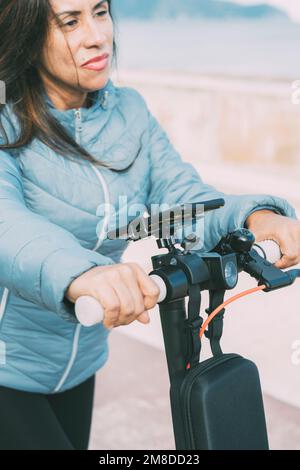 Wunderschöne Frau, die mit ihrem Motorroller in der Stadt entlang der Uferpromenade fährt, umweltfreundliche Lokomotionsfahrzeuge und umweltfreundliche ec Stockfoto