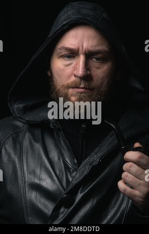 Porträt eines bärtigen Mannes in schwarzer Regenjacke, der eine Pfeife raucht Stockfoto