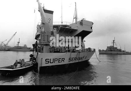 Ein Arbeitsboot bewegt sich entlang des Aufbaus des holländischen Schwertransportschiff SUPER SERVANT 3, während sich der Meeresminenweeper USS LEADER (MSO-490) nähert, um an Bord des Unterdecks des Schiffes zu laden. Der SUPERDIENER 3 wird als Reaktion auf die irakische Invasion Kuwaits den FÜHRER, andere Minenräumer und das Mine-Gegenschiff USS AVENGER (MCM-1) in den Persischen Golf transportieren. Betreff Betrieb/Serie: DESERT STORMDESERT SHIELD Base: Naval Air Station, Norfolk Bundesstaat: Virginia (VA) Land: Vereinigte Staaten von Amerika (USA) Stockfoto