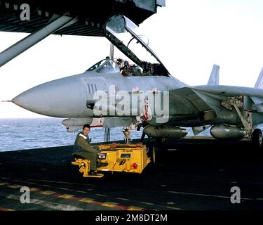 Ein Besatzungsmitglied des Cockpits steht mit einem SD-1D bereit, der neben einem Fighter Squadron 14 (VF-14) F-14A Tomcat-Flugzeug einen Dolly beobachtet, während das Flugzeug von einem abgesenkten Aufzug an Bord des Flugzeugträgers USS JOHN F. KENNEDY (CV 67) zum Abschleppen vorbereitet ist. Basis: USS John F. Kennedy (CV 67) Stockfoto