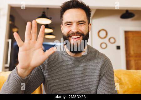 Porträt eines glücklichen birassischen jungen Mannes, der Hand winkt, während er im Wohnzimmer auf dem Sofa sitzt Stockfoto