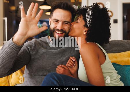 Ein bärtiger birassischer Mann winkte mit der Hand und eine Freundin küsste ihn auf der Wange, während sie sich auf dem Sofa entspannte Stockfoto