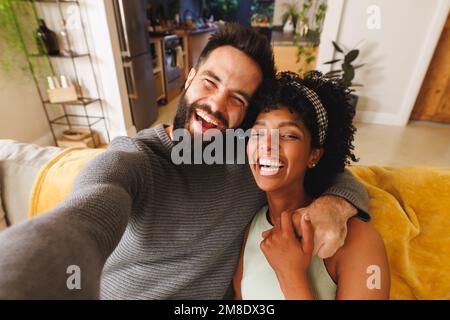 Porträt eines glücklichen birassischen bärtigen Mannes, der die Hände seiner Freundin hält und Selfie auf dem Sofa macht Stockfoto