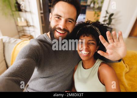 Porträt eines glücklichen birassischen bärtigen Mannes, der Hand winkt und Selfie mit seiner Freundin auf dem Sofa macht Stockfoto