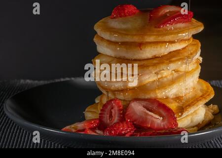 Frisch geschnittene Erdbeeren mit einem Stapel Buttermilch-Pfannkuchen, getränkt mit Honig auf einem dunklen Teller mit dunklem Hintergrund und viel Negativraum Stockfoto