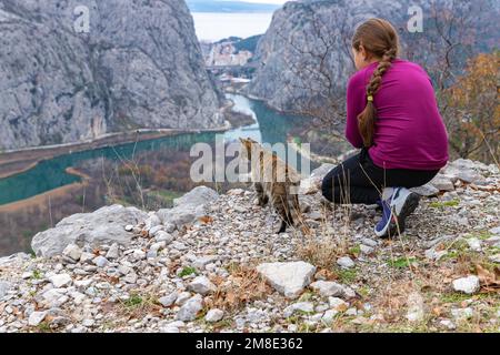 Mädchen und Katze sitzen auf dem Gipfel des Berges und schauen auf den Fluss Cetina und die Adria nahe Omiš in Kroatien Stockfoto