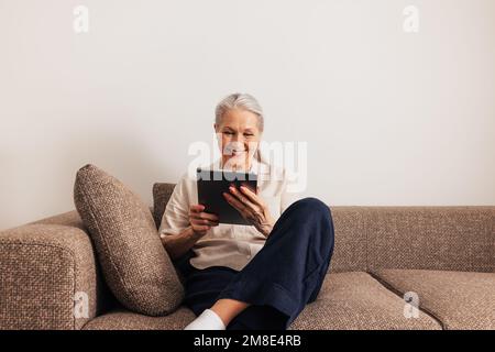 Fröhliche Erwachsene Frau, die auf einem Sofa mit einem digitalen Tablet sitzt. Lächelnde Seniorin mit einem tragbaren Computer im Wohnzimmer. Stockfoto