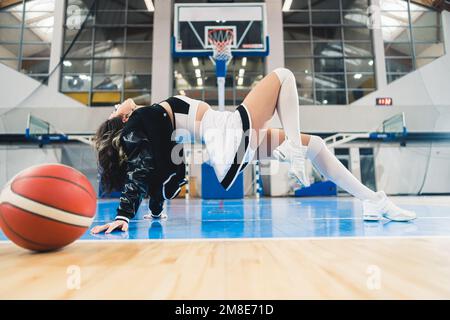 Der Cheerleader zeigt in der sportlichen Pose Leichtfertigkeit. Ein Basketball wird in der Nähe platziert. Hochwertiges Foto Stockfoto