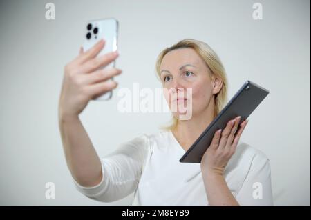 Eine Erwachsene Frau macht ein Selfie mit einem Telefon in der anderen Hand. Sie hat ein Tablet mit weißem Hintergrund, weißes T-Shirt, offene Augen machen die Gesichter mit einem weißen Telefon gestreckt Stockfoto