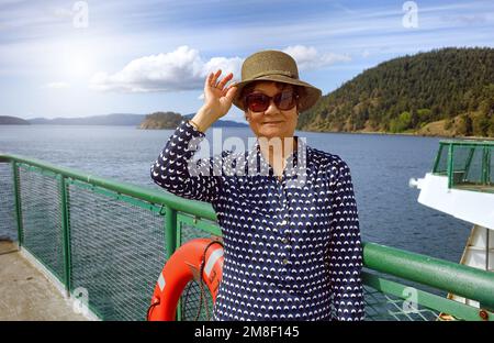 Seniorin, die sich nach vorne freut, Hut und Sonnenbrille trägt, während sie auf dem Bootsanleger steht, mit Meer und hellem Tageslicht im Hintergrund Stockfoto