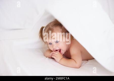 Ich verstecke mich vor mom. Ein süßes kleines Mädchen, das sich unter den Bettdecken versteckt. Stockfoto