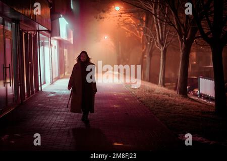 Eine einsame junge Frau im Mantel läuft durch die nächtliche neblige Stadt der Felder, die von Lampen beleuchtet werden Stockfoto