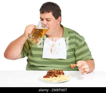 Fest mit Fett. Ein fettleibiger junger Mann, der ein Lätzchen trägt und ein Bier trinkt, mit einem Teller Hühnerflügel und Pommes Frites vor ihm. Stockfoto