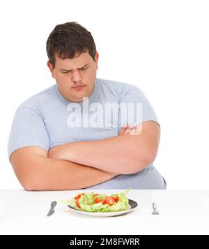 Niemand hat Zeit für Salat. Ein fettleibiger junger Mann, der mit gefalteten Armen an einem Tisch sitzt und einen Teller Salat vor sich hat. Stockfoto