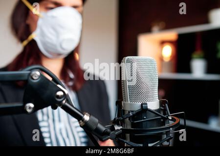 Frau mit Gesichtsmaske sendet ihren Podcast mit einem Mikrofon aus ihrem Studio. Stockfoto