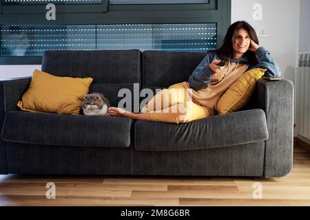Entspannte Frau, die lächelt, während sie fernsieht, während ihre Katze auf dem Sofa sitzt. Stockfoto