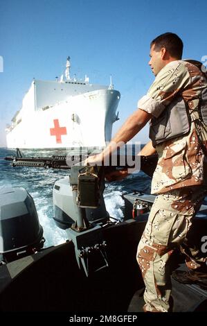 EIN US-AMERIKANISCHER Küstenwache, ein Mitglied der Hafensicherheit patrouilliert während der Operation Desert Shield in einem Jägerboot mit M-60 Maschinengewehr. Im Hintergrund befindet sich das Spitalschiff USNS MERCY (T-AH-19) des militärischen Sealift-Kommandos. Betroffene Operation/Serie: WÜSTENSCHILD Land: Unbekannt Stockfoto