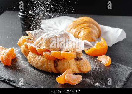 Croissant mit süßen Mandarinen auf dunklem Hintergrund. Hausgemachte Croissants. Fallender Puderzucker. Vertikal. Stockfoto