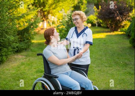 Eine ältere Krankenschwester geht mit einer Frau mittleren Alters im Rollstuhl im Park. Stockfoto