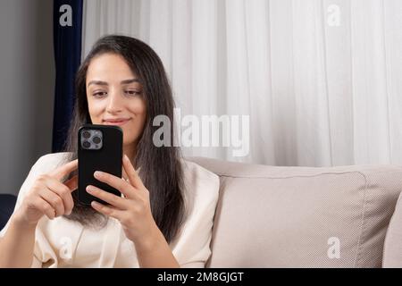 Mit dem Smartphone, einer weißen Frau, die modern ist und ein Smartphone benutzt. Zu Hause sitzen, lächeln, mit einem Freund chatten oder im Internet surfen, soziale Medien nutzen. Stockfoto