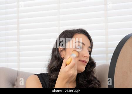 Sie trägt Rouge auf, braune Millennialfrau. Beigefarbenes Fundamentpulver auf den Wangen. Mit Make-up-Pinsel. Sessel zu Hause. Make-up-Konzept. Stockfoto