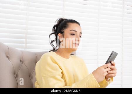 Frau, die zu Hause sitzt und ein Smartphone benutzt. Kaukasisches, brünettes Mädchen der Millennials, das auf dem Handy zusieht, chattet, soziale Medien nutzt. Konzept der Technologie. Stockfoto