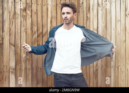 Ein junger gutaussehender Mann steht auf einem hellen Holzhintergrund. Der Kerl trägt ein weißes, leeres T-Shirt ohne Logo, was es perfekt sui macht Stockfoto