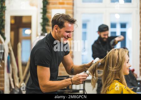 Freudiger Friseur trocknet und kämmt blondes Frauenhaar, ein weiterer Friseur arbeitet im Hintergrund. Hochwertiges Foto Stockfoto