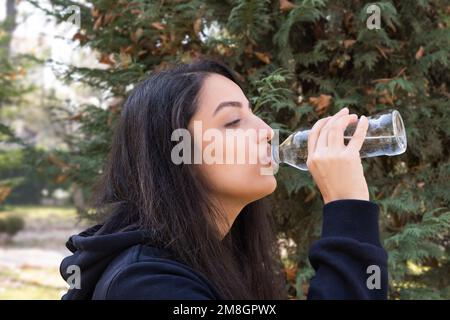 Trinkwasser aus Glasflasche. Seitenansicht einer Frau, die reines Mineralwasser trinkt. Im Park. Lässige Sportkleidung im Herbst. Gesunde Gehgewohnheit. Stockfoto