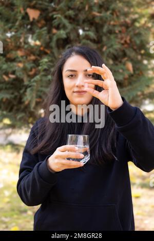 Eine Frau, die die Pille in der Hand hält. Frontansicht Porträt der Frau zeigt Tablet, Medizin, Gesundheitskonzept. Die Kamera steht draußen. Ein Glas Wasser Stockfoto
